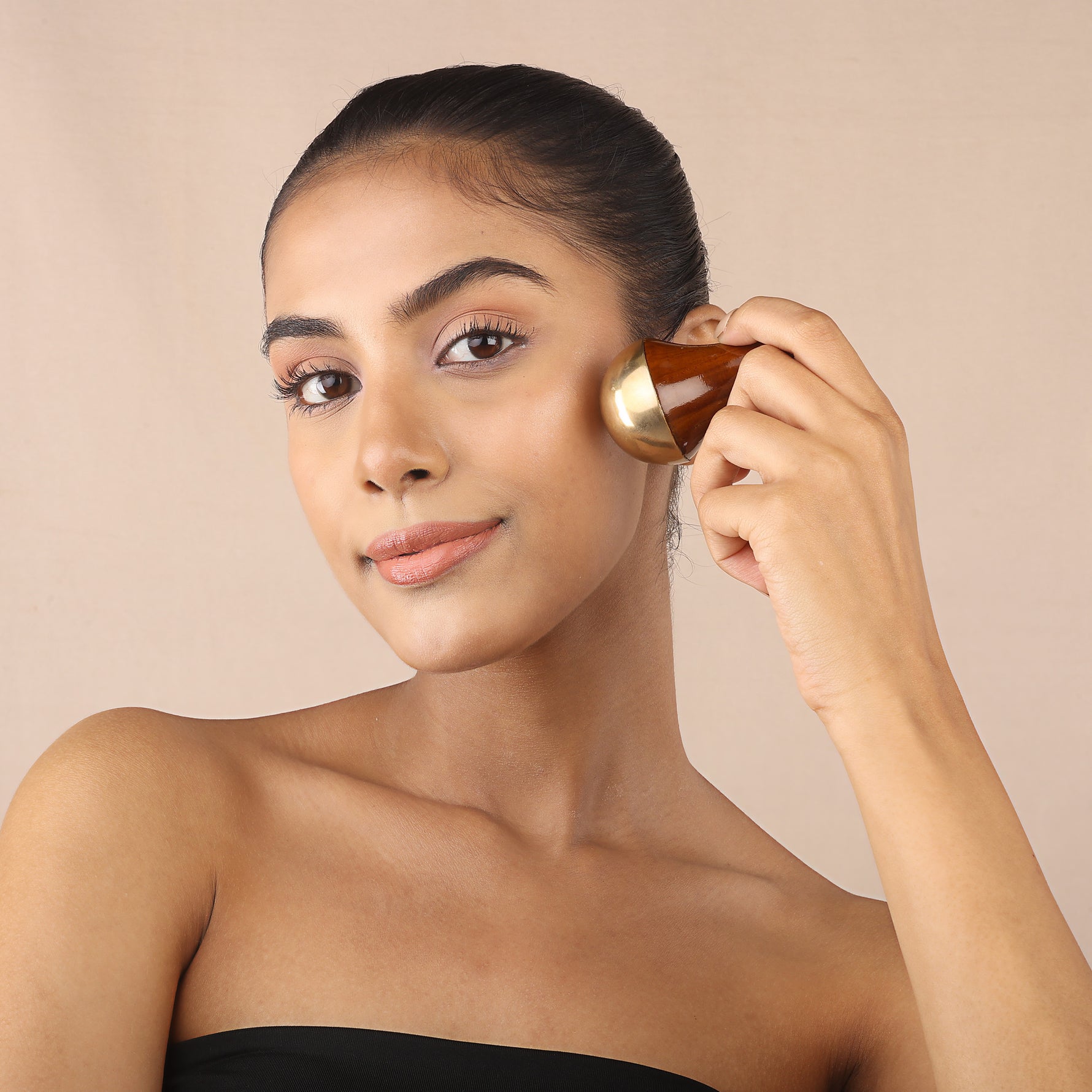 Woman using an Art of Vedas Kansa face wand on her cheek as part of the Ayurveda Kansa facial ritual against a warm neutral background