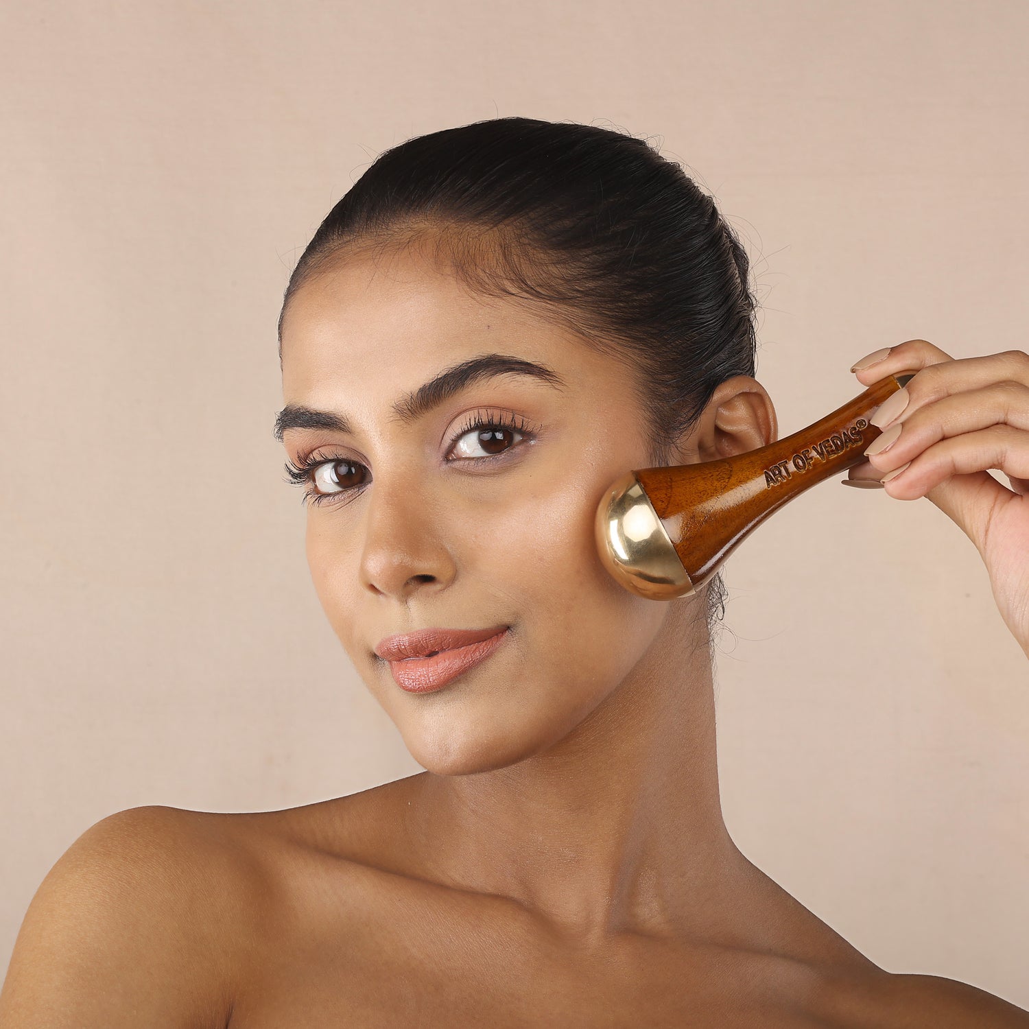 Close-up of a woman holding an Art of Vedas Kansa metal face wand near her cheek, showcasing the bronze and wood facial massage tool against a warm neutral background