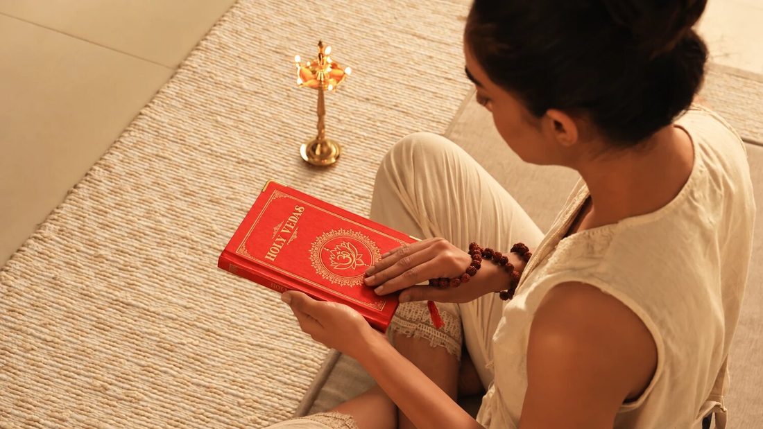 Kvinde, der sidder på en vævet måtte og læser den røde bog Holy Vedas ved siden af en ayurvedisk messinglampe, for Art of Vedas